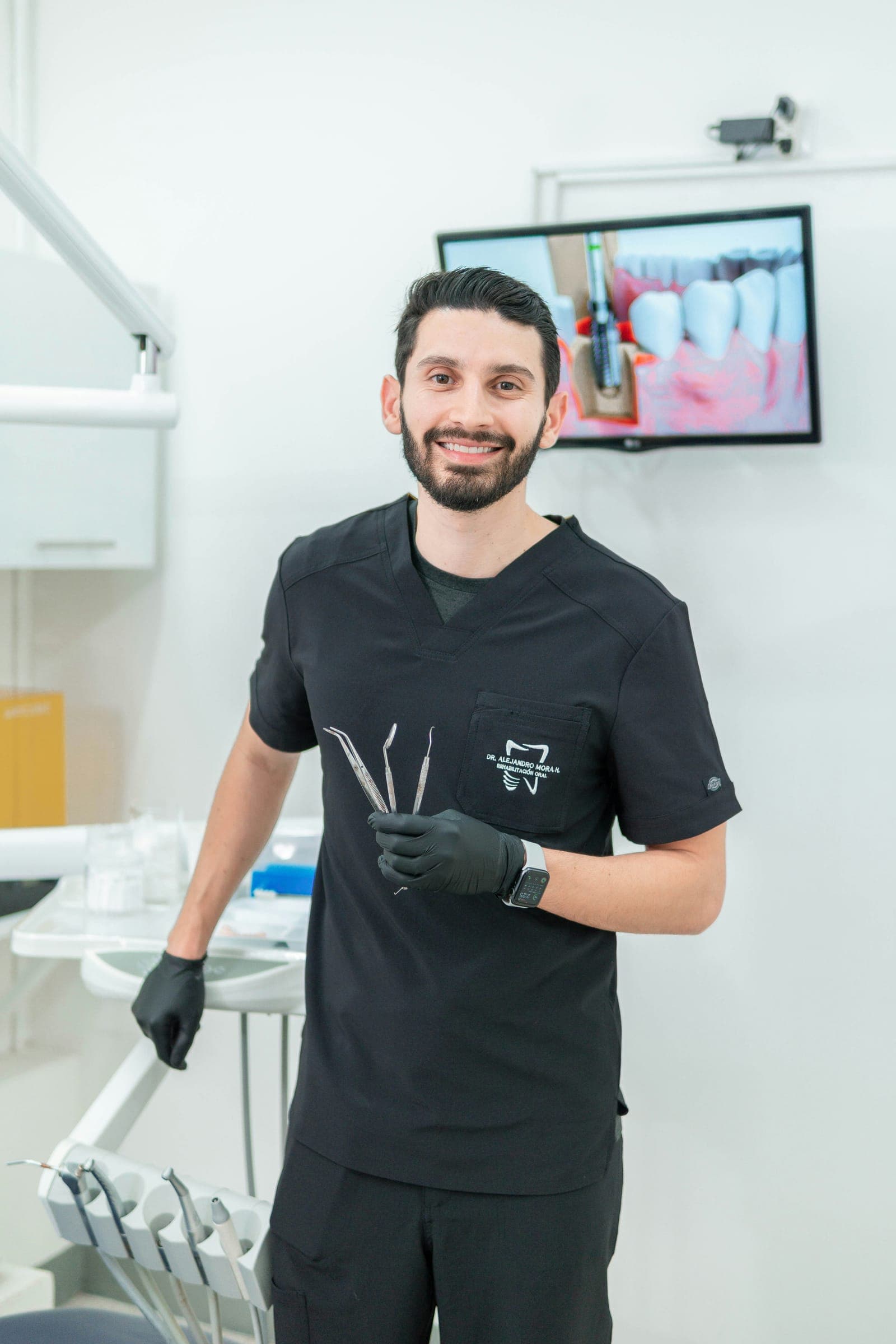 Dentista de uniforme preto sorrindo no consultório, com instrumentos clínicos nas mãos.