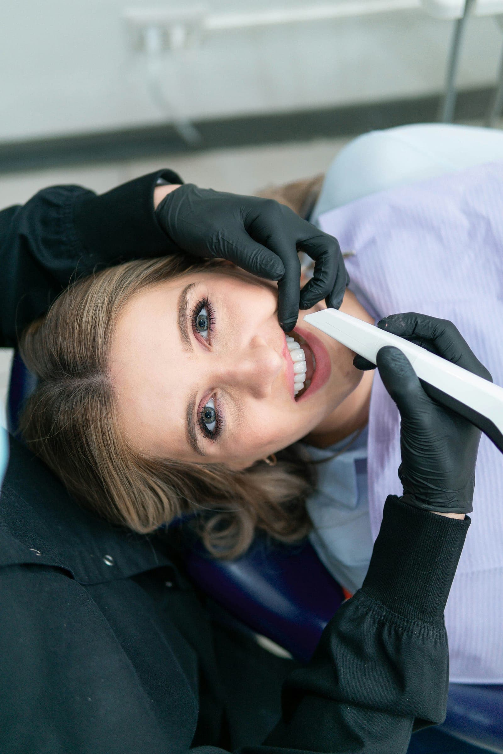 Paciente sorri na cadeira enquanto o dentista, de luvas pretas, faz o escaneamento intraoral.