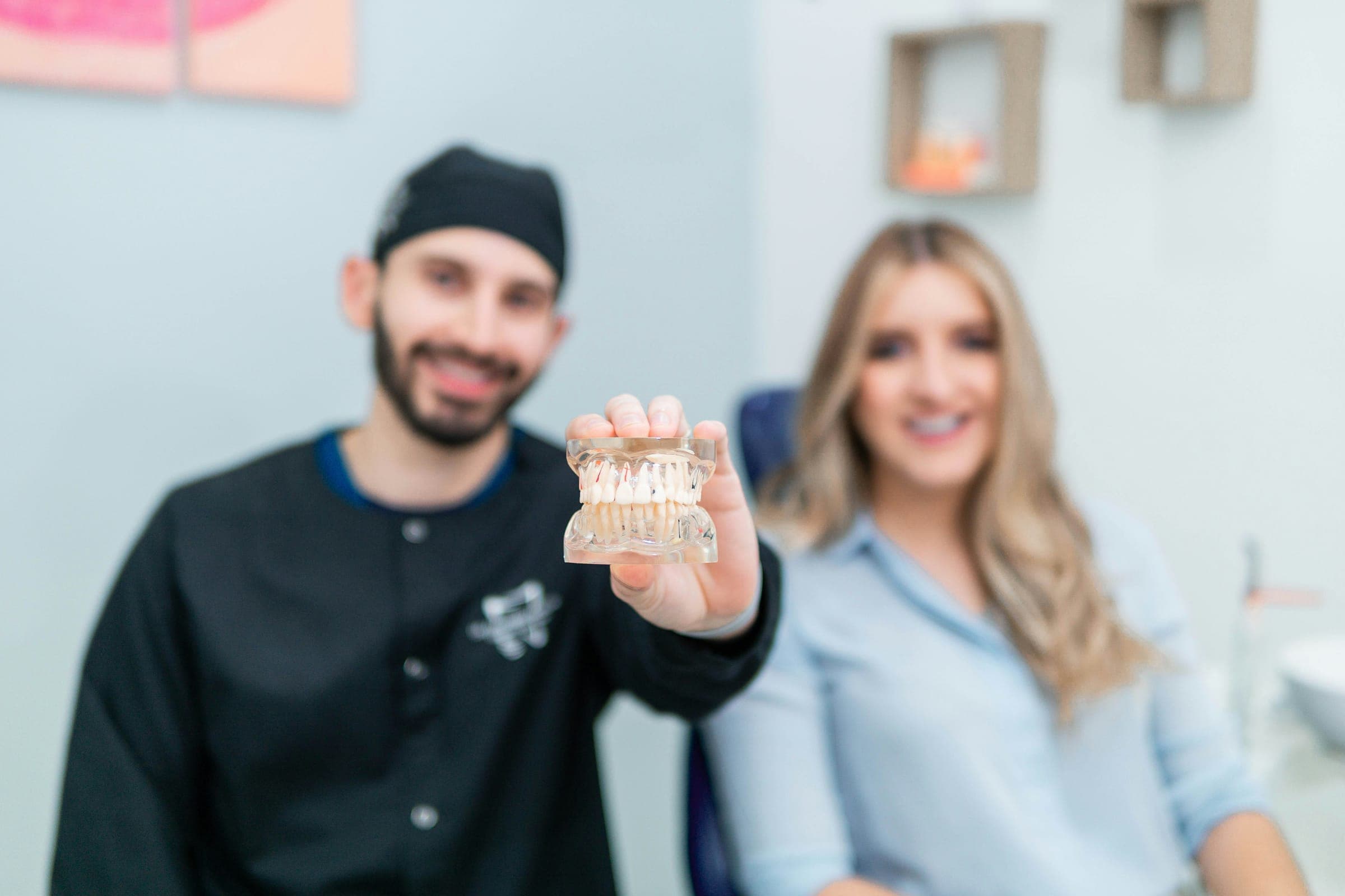 Dentista segura um modelo de arcada dentária; ao fundo, uma paciente sorri.